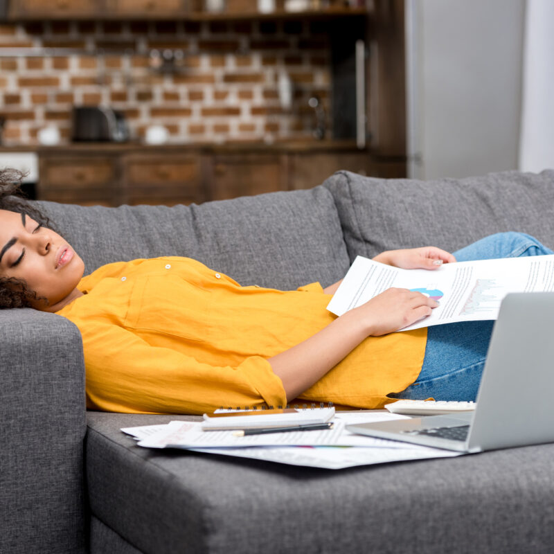 A tired Black woman reclines on her couch with papers spread around her.