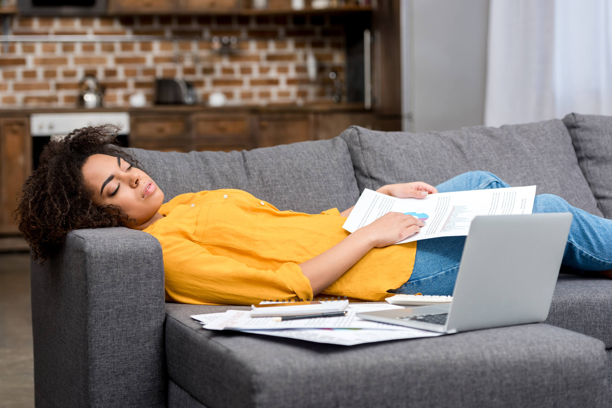 A tired Black woman reclines on her couch with papers spread around her.