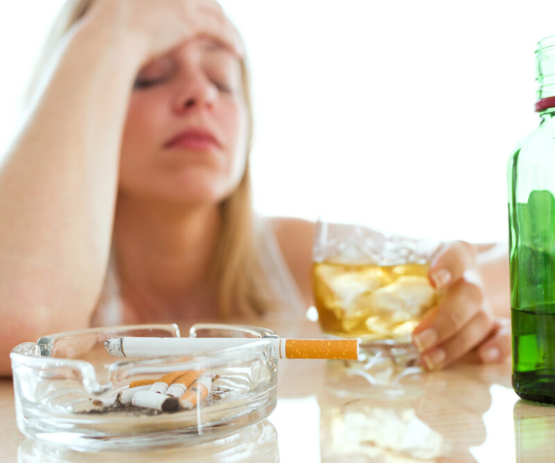 woman holding her head in her hand and holding a glass of whiskey with the other, cigarettes on table in front of her