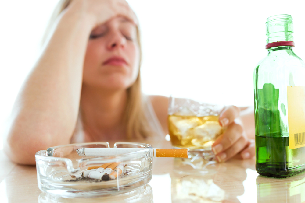 woman holding her head in her hand and holding a glass of whiskey with the other, cigarettes on table in front of her