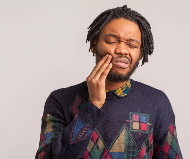 Frustrated bearded african man with dreadlocks touching cheek and wincing in pain feeling terrible tooth ache, popping jaw, gingivitis, periodontal disease. Indoor studio shot isolated on gray background