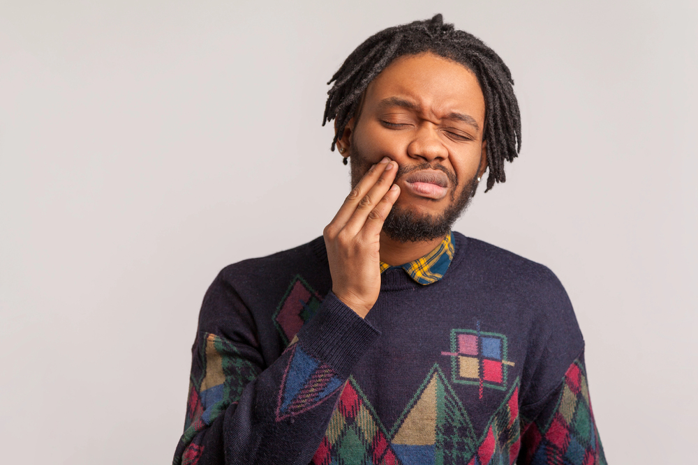 Frustrated bearded african man with dreadlocks touching cheek and wincing in pain feeling terrible tooth ache, popping jaw, gingivitis, periodontal disease. Indoor studio shot isolated on gray background