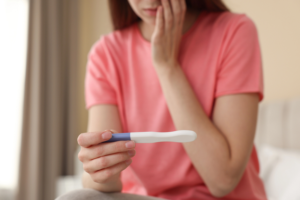 woman in pink shirt sitting on bed looking at pregnancy test result